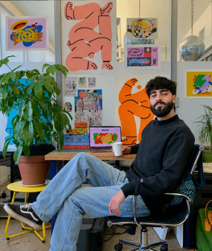 Caio Martins sentado na mesa de trabalho, com cartazes ilustrados na parede ao fundo